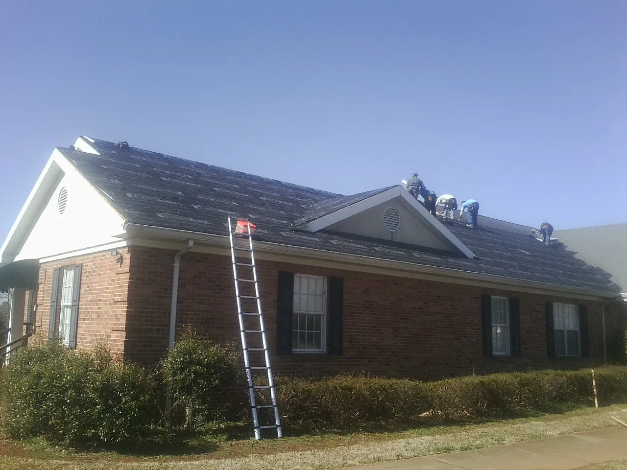 Crew tearing shingles off a brick ranch roof with a ladder set — Metal Roof Repair work by our crew in Howe